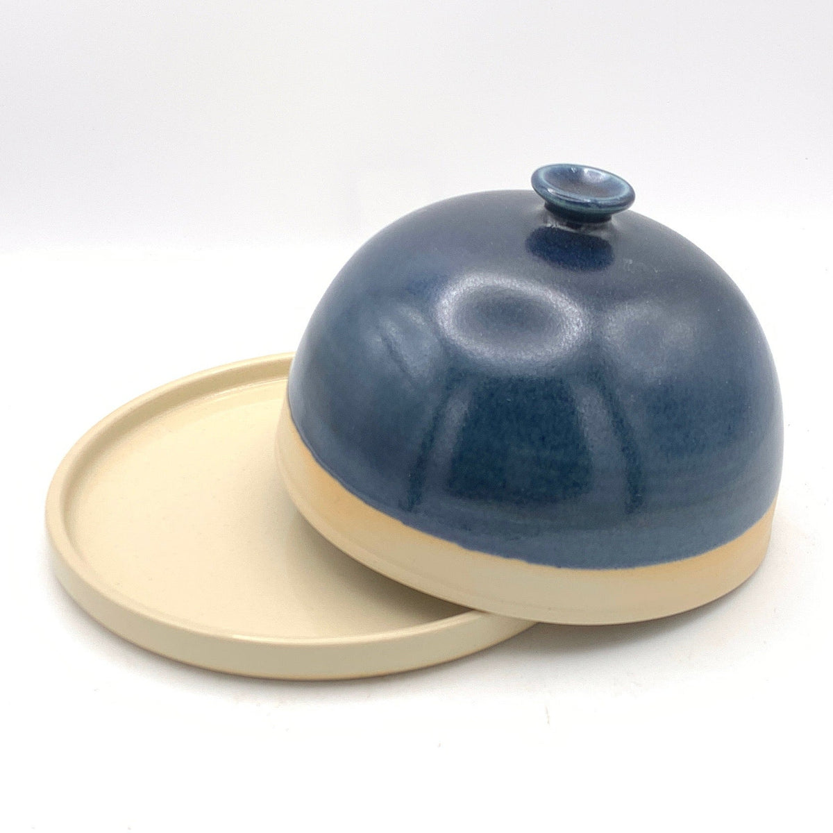 A small ceramic cloche or dome-shaped container with a matching round lid, shown inside a white lightbox. The cloche has a glossy dark blue glazed top and a matte beige base, while the lid is plain matte beige.