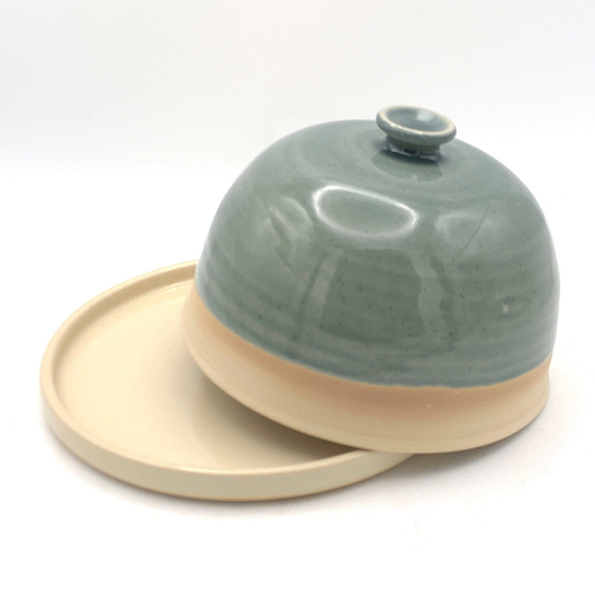 A small ceramic lidded dish or bowl with a pale green glazed top and a natural beige unglazed base, shown on a white studio background with a black fabric surround.