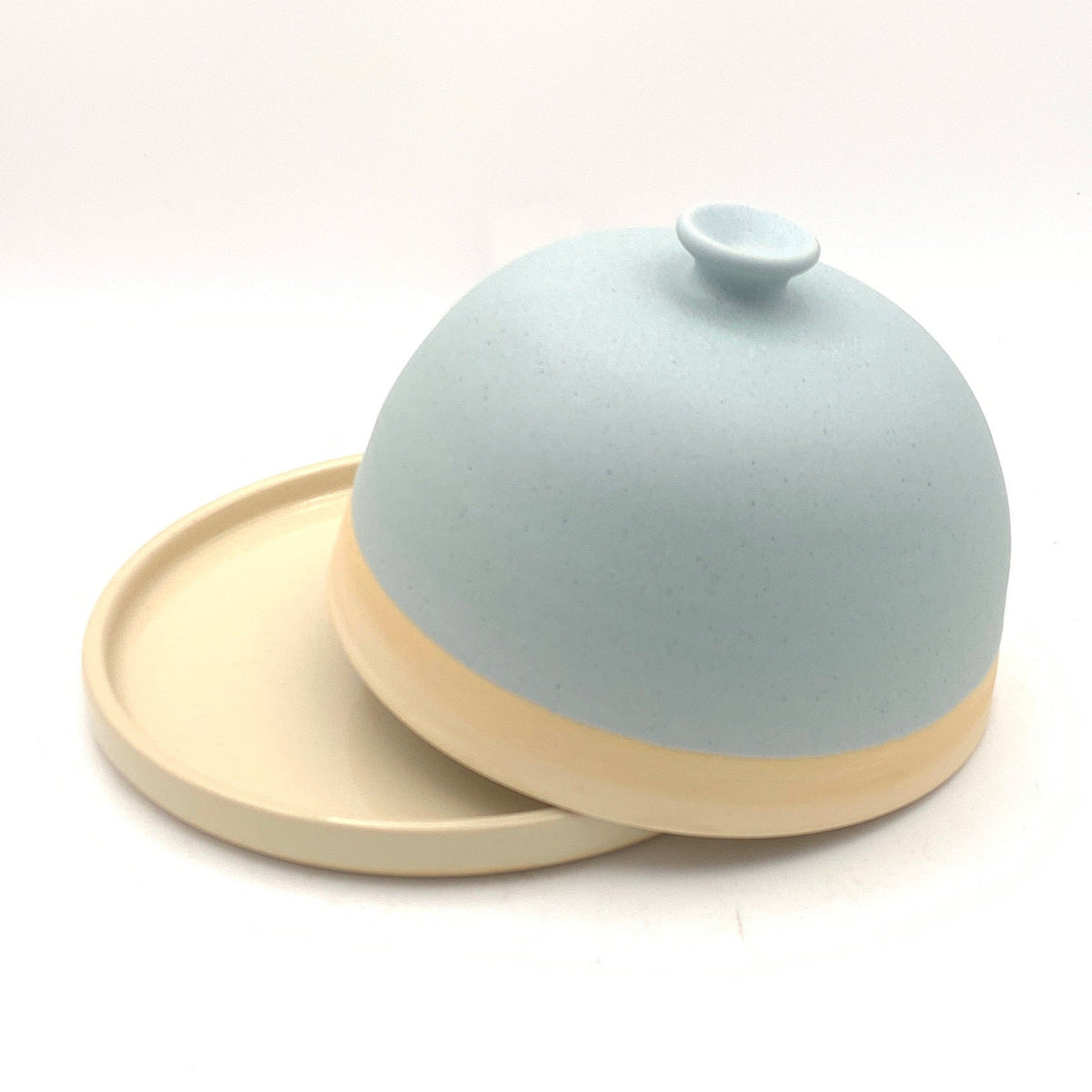 A small dome-shaped ceramic container with a matching round base, shown inside a lightbox. The container has a matte pale blue top and a warm beige base, with the lid resting on the base.