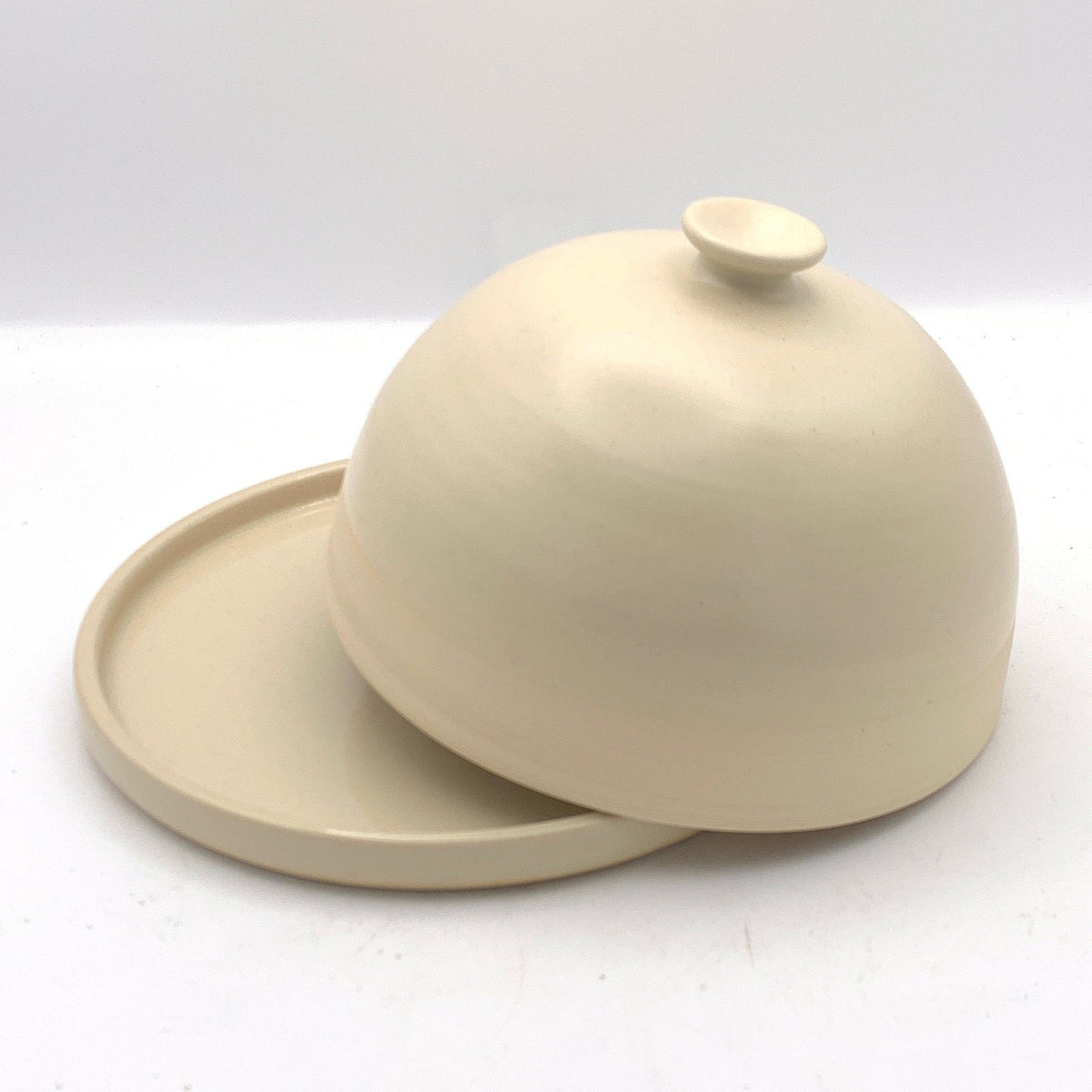 A small ceramic lidded dish or covered plate in a warm cream or off-white glaze, photographed inside a white light box with a black foreground band at the bottom.