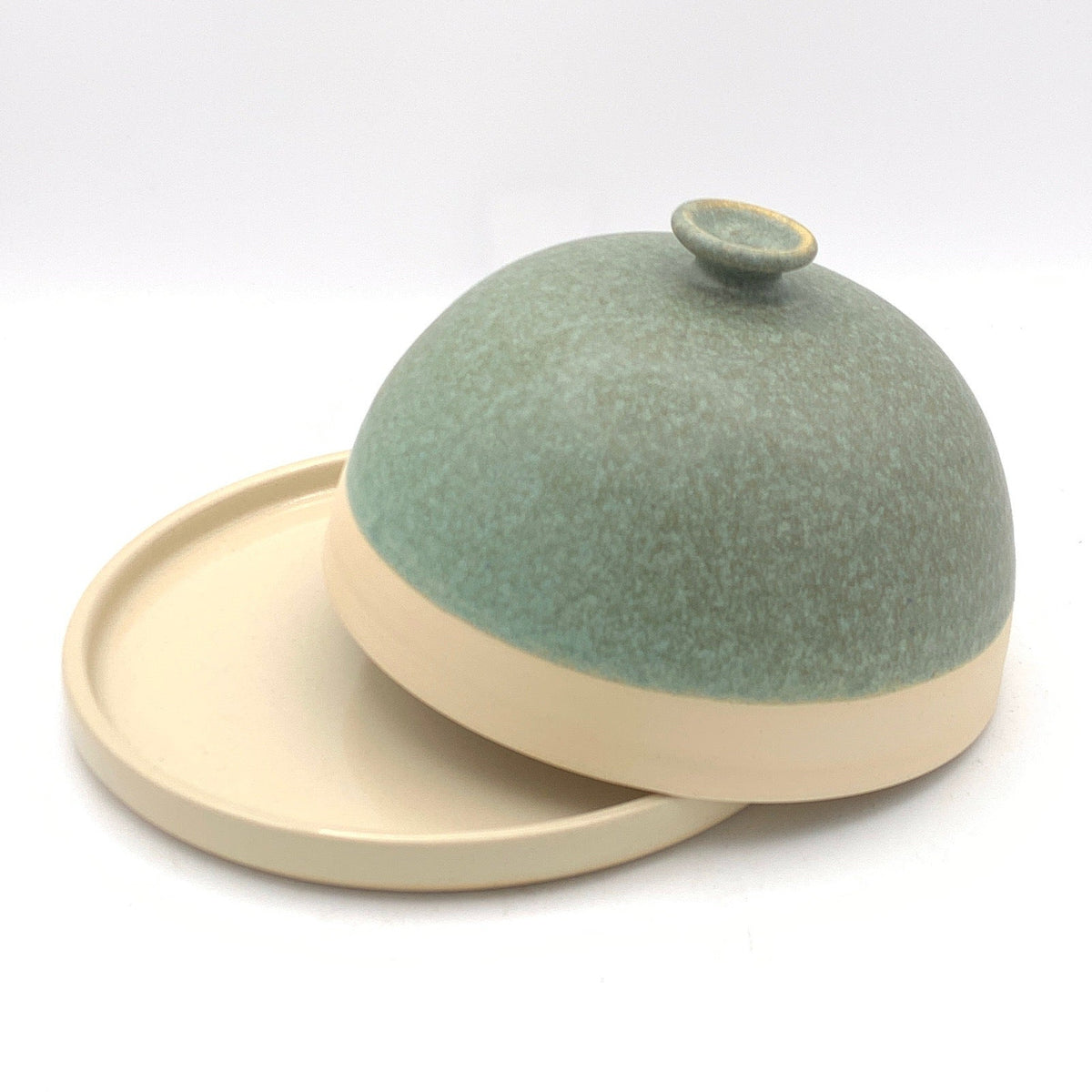 A small ceramic dome-shaped container with a matching round base and lid, shown inside a lightbox. The container has a speckled sage green top half and a matte cream-colored bottom half and lid.