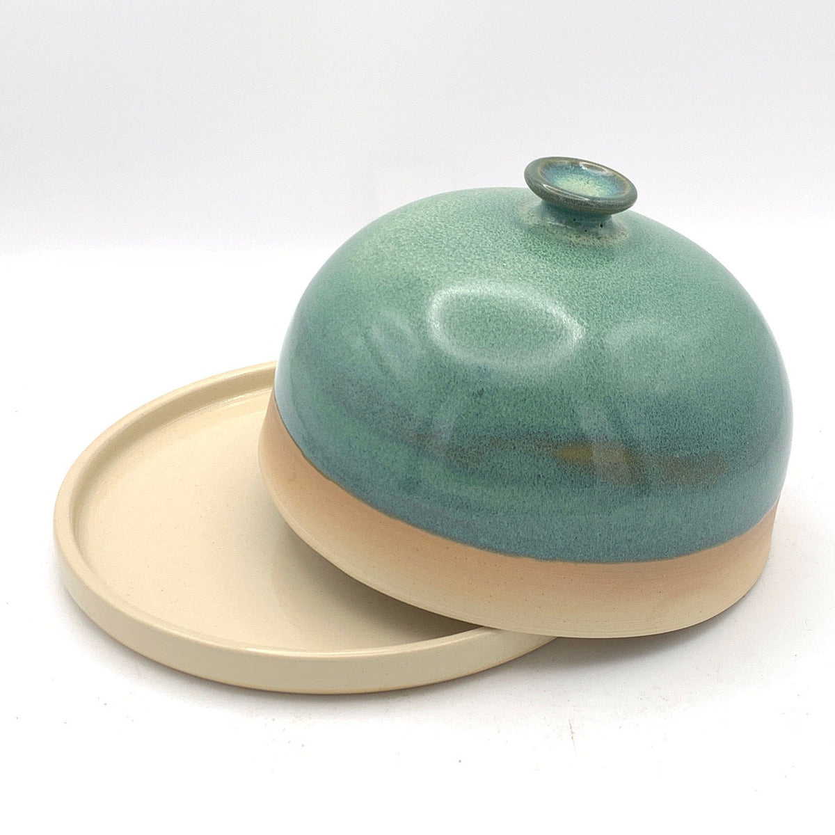 A small ceramic lidded dish or covered plate with a rounded dome-shaped top in a glossy teal-green glaze and a beige base, shown on a white studio background with a black fabric surface at the bottom.
